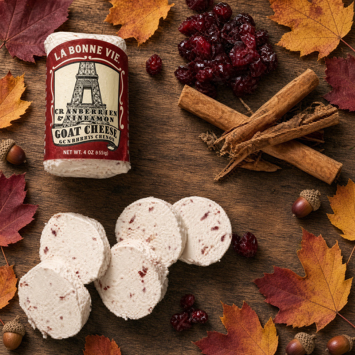 A wheel of La Bonne Vie Cranberry & Lemon Goat Cheese is shown on a rustic wooden surface with sliced cheese pieces, dried cranberries, cinnamon sticks, acorns, and autumn leaves. The cheese label is clearly visible, and the food is styled in a seasonal, appetizing arrangement.