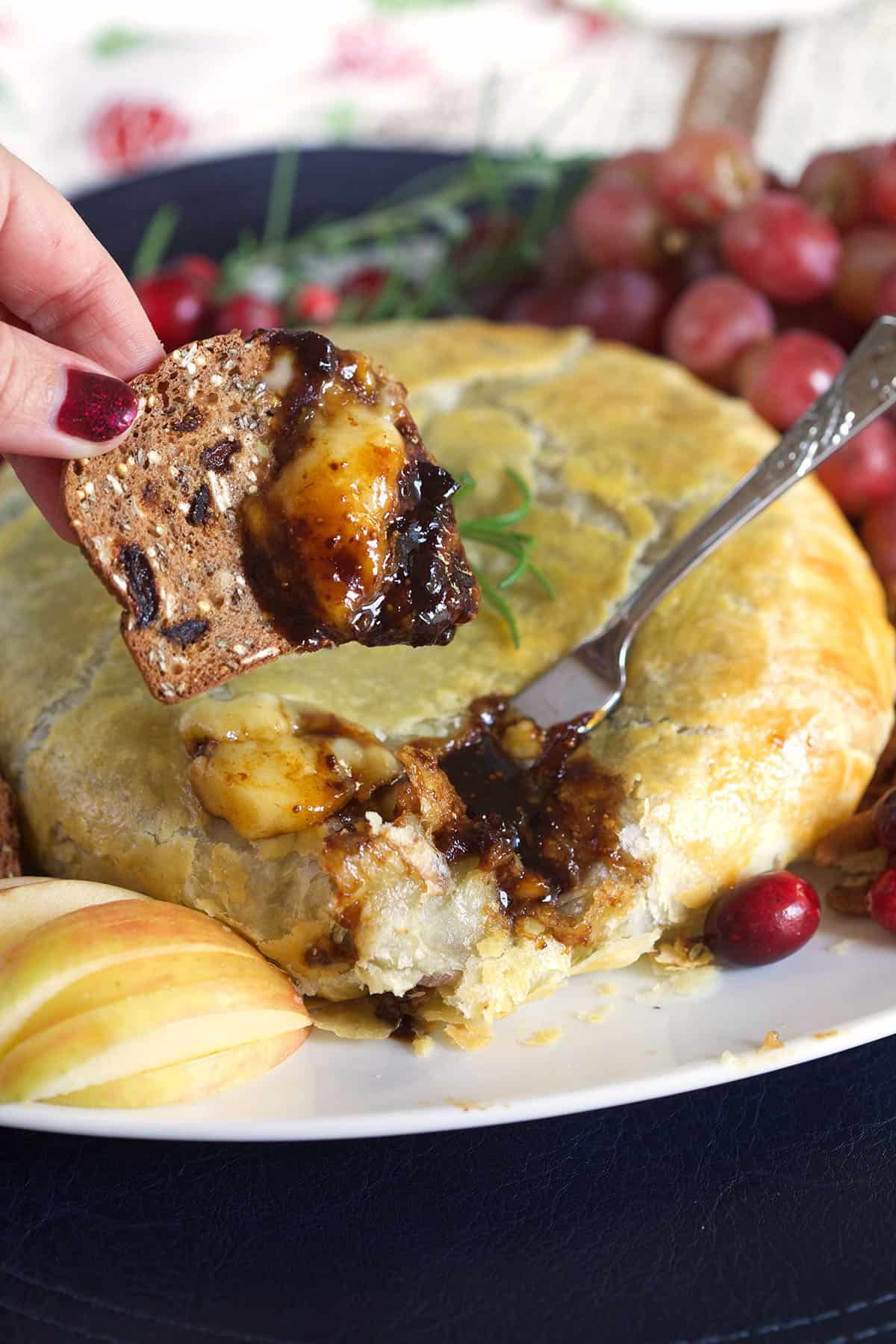 A close-up of a baked cheese pastry or cheese bread on a white plate, with a hand dipping a cracker into melted cheese and sauce. A fork rests in the pastry, and cranberries, grapes, and apple slices are arranged around it on a dark table.