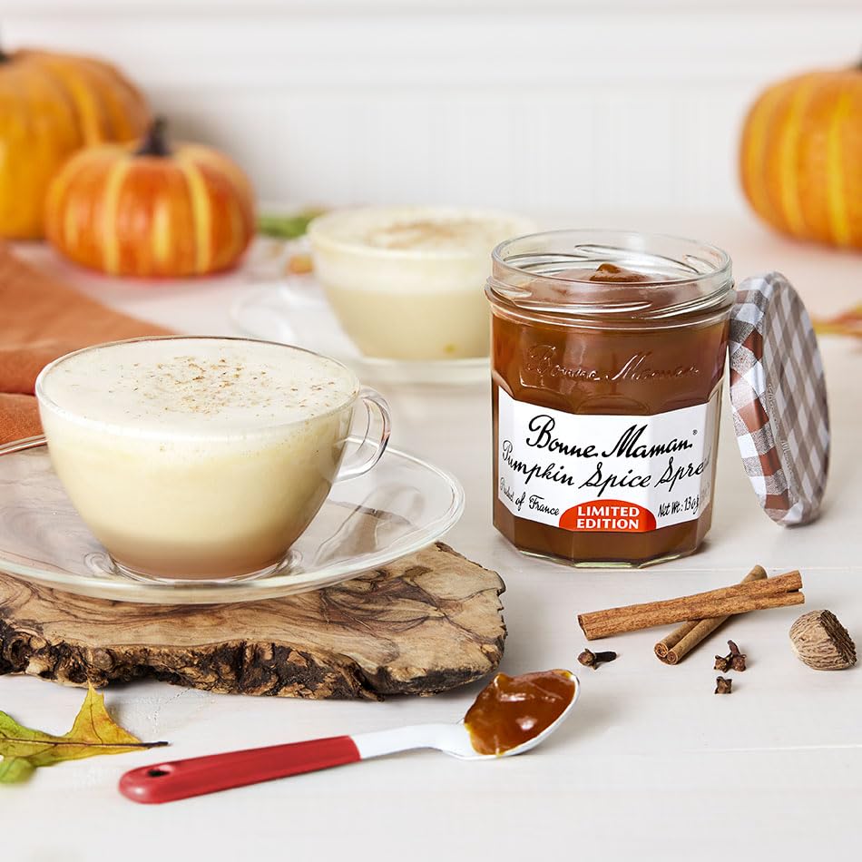 A jar of Bonne Maman Pumpkin Spice Spice spread sits on a white surface beside a frothy latte, cinnamon sticks, a spoon with spread, and small pumpkins in the background.