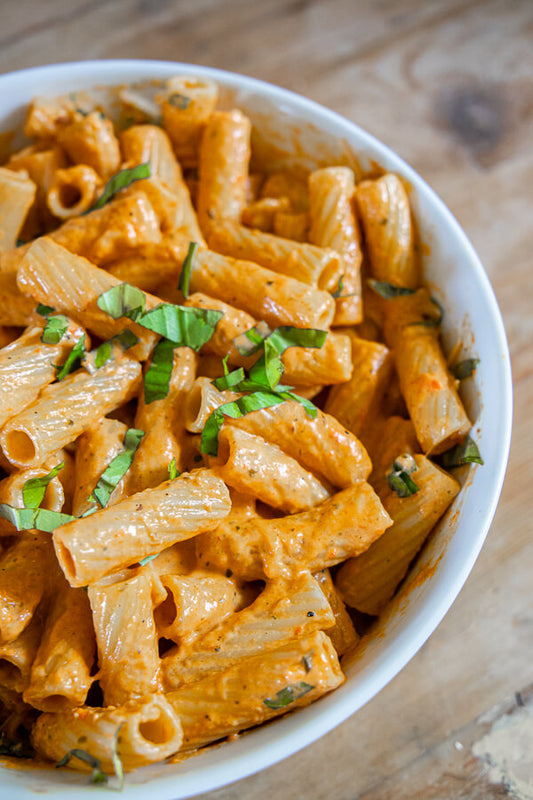 A close-up shot of a white bowl filled with penne pasta in a creamy orange-red sauce, garnished with chopped green herbs, set on a rustic wooden surface.