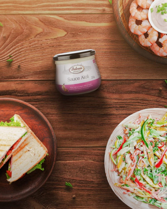 A jar of Sabreleau Sauce Ailini placed on a rustic wooden table, surrounded by food props including a sandwich on a plate, a shrimp dish, and a salad.
