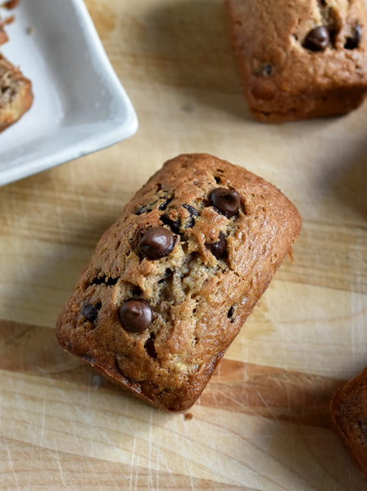 Close-up of a My Big Fat Cookie mini banana chocolate chip bread loaf on a wooden cutting board, showing golden-brown crust studded with melted chocolate chips, with more mini loaves in the background