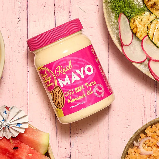 A jar of Real Mayo almond butter with a bright pink lid and label, styled on a pink wooden surface with sliced radishes, watermelon, and grilled zucchini around it.