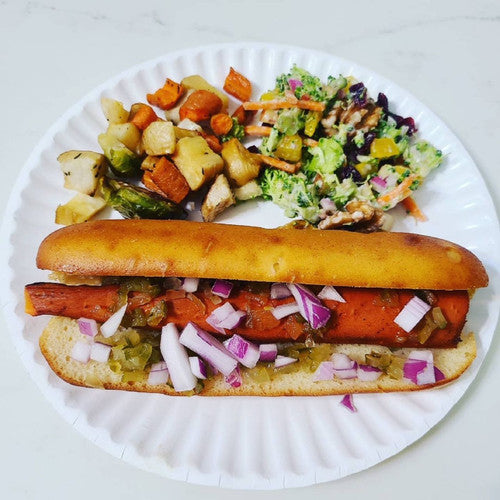 Gluten free hot dog bun topped with red onion, relish, and peppers, served on a paper plate with roasted vegetables and coleslaw