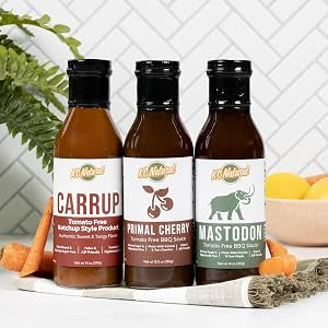 Three bottles of 10 Natural spicy buffalo sauce arranged on a white surface with a tiled wall behind. The bottles are labeled Carrup, Primal Cherry, and Mastoon, with carrots, herbs, and lemons used as styling props.