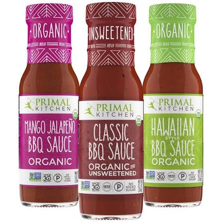 Three Primal Kitchen Organic BBQ Sauce bottles in a row: Mango Jalapeño, Classic Unsweetened, and Hawaiian Style. The bottles show the dark reddish-brown sauce inside, with colorful labels and caps in pink, white, and green.