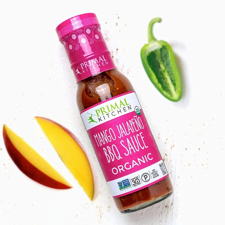 Bottle of Primal Kitchen Mango Jalapeño BBQ Sauce with a slice of mango and jalapeño on a white background
