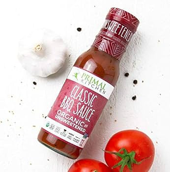 A bottle of Primal Kitchen Classic BBQ Sauce, Organic Unsweetened, styled on a white textured background with garlic, tomatoes, and black pepper.