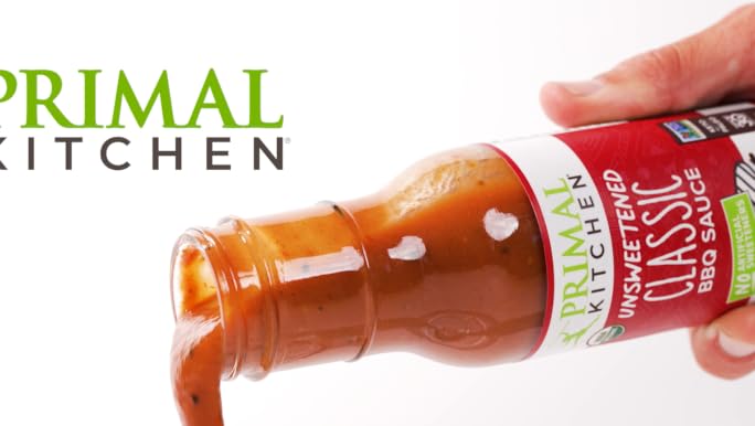 A hand holding a bottle of Primal Kitchen Classic Unsweetened BBQ Sauce, with sauce pouring out, on a white background with the Primal Kitchen logo in the upper left.