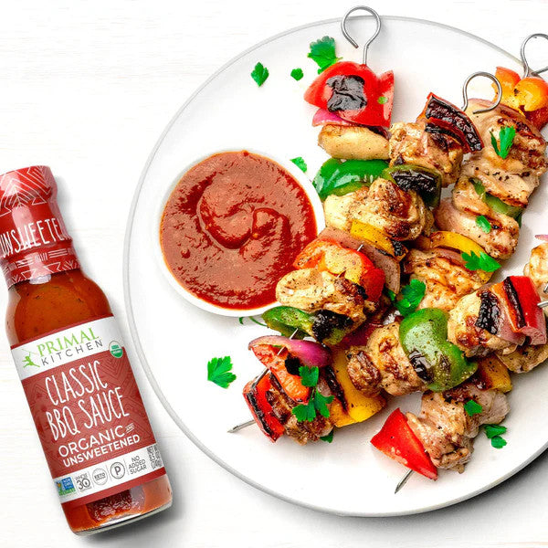 A bottle of Primal Kitchen Classic BBQ Sauce next to a white plate with grilled chicken and vegetable skewers and a small bowl of the red sauce on a white background.