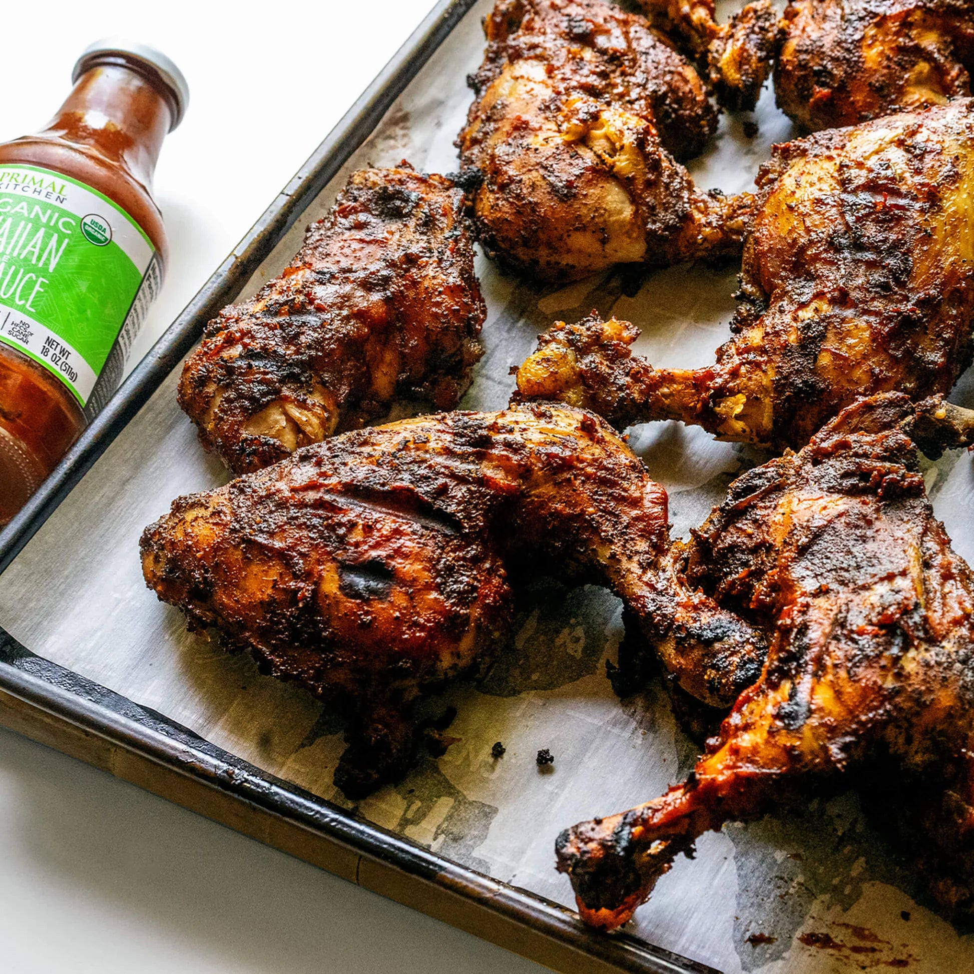 Spiced chicken legs on a baking tray with a bottle of hot sauce.