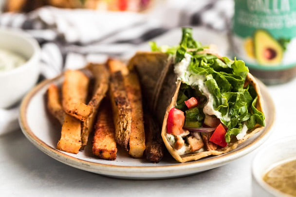 A close-up of a plated gourmet wrap filled with chicken, greens, tomatoes, and a white dressing, served with thick roasted sweet potato fries on a ceramic plate. A bottle of avocado dressing and small dipping bowls are visible in the softly blurred background.