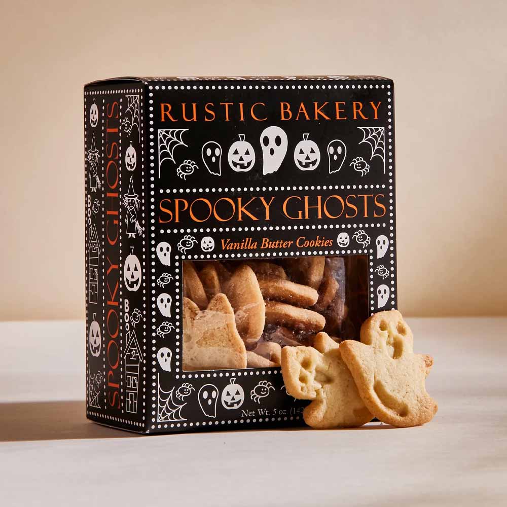 Rustic Bakery Spooky Ghosts vanilla butter cookies in a black Halloween-themed box with ghost and pumpkin illustrations, with two ghost-shaped cookies placed in front on a beige surface.