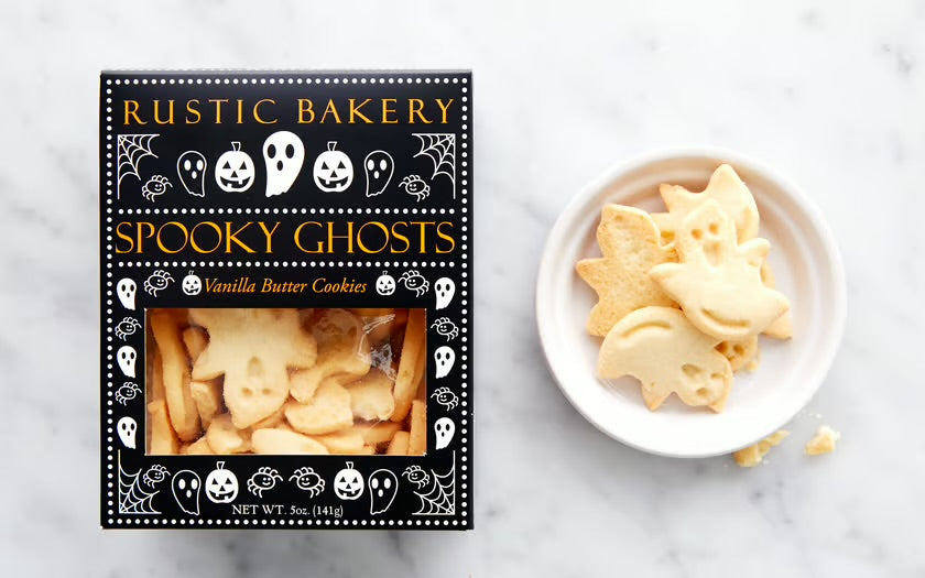 Rustic Bakery Spooky Ghosts vanilla butter cookies in a black decorative box next to a white dish of ghost-shaped cookies on a light marble surface.