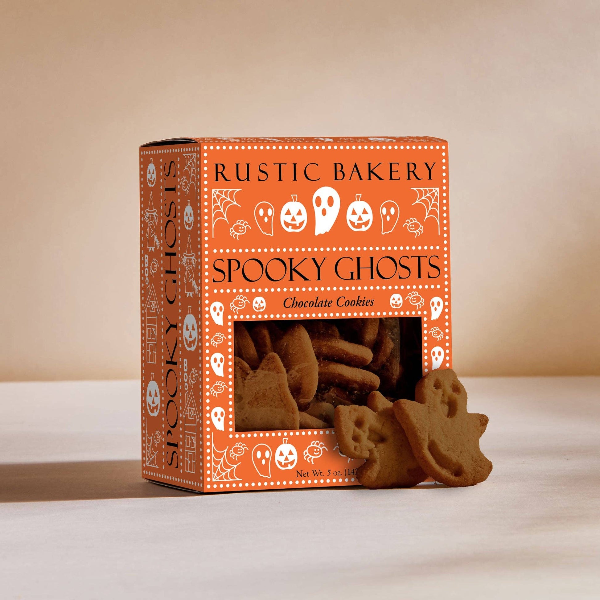 A box of Rustic Bakery Spooky Ghosts chocolate cookies on a light beige surface with a matching background. The orange packaging features white Halloween-themed illustrations, and several ghost-shaped cookies are displayed in front of the window.