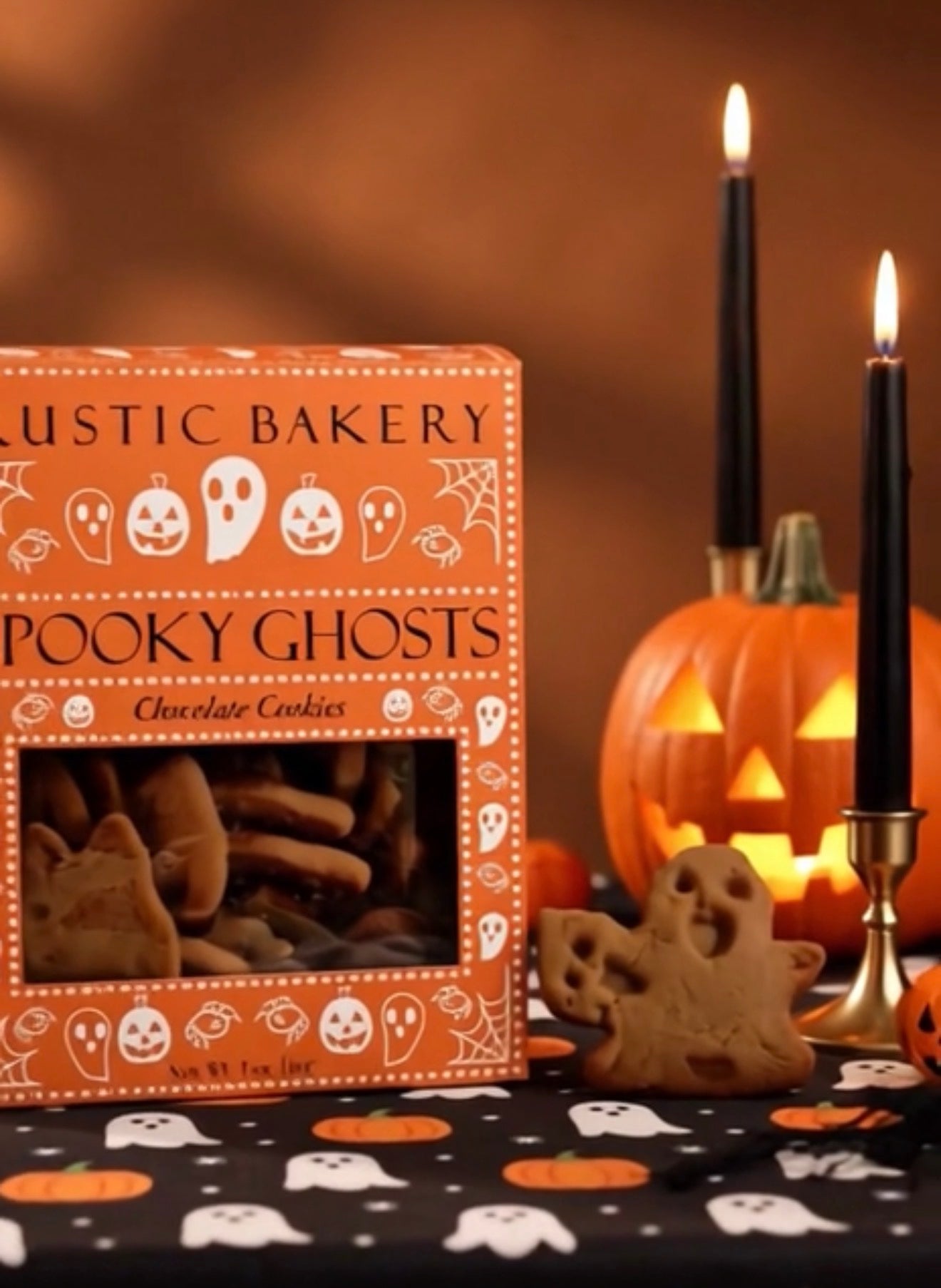 A box of Rustic Bakery 'Spooky Ghosts' chocolate cookies displayed on a Halloween-themed tablecloth with black candles, a carved pumpkin, and a small ghost cookie in front.