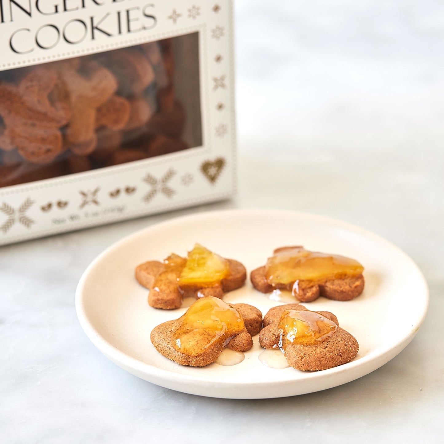 Four gingerbread person-shaped cookies topped with amber syrup on a small white plate, with a box of gingerbread cookies blurred in the background.