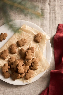 Gingerbread cookies shaped like small figures, arranged on parchment paper over a white plate on a linen surface, with a red cloth and blurred greenery in the background.