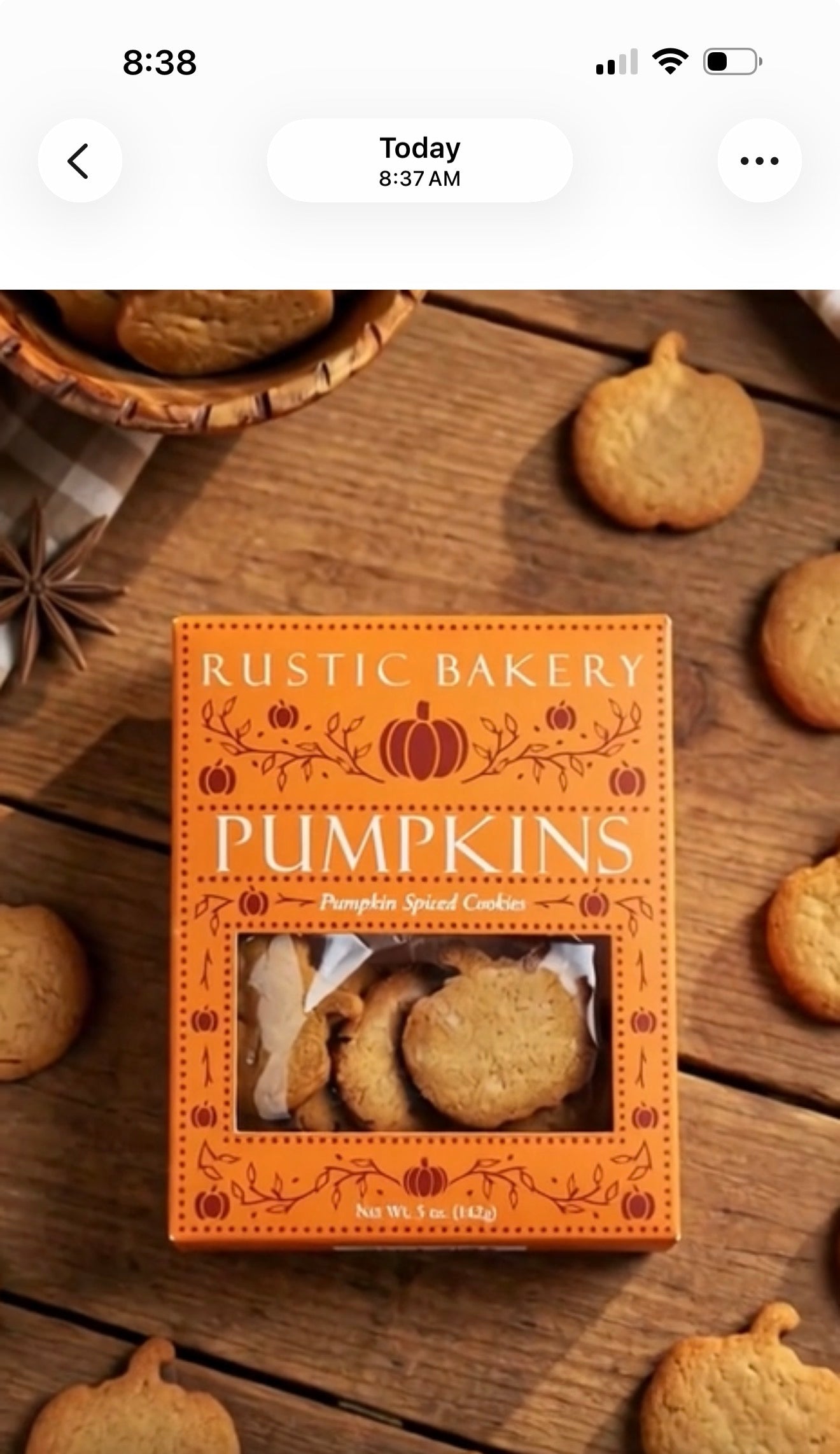 A box of Rustic Bakery Pumpkins cookies placed on a rustic wooden table, surrounded by scattered pumpkin-shaped cookies and a basket of cookies in the background.