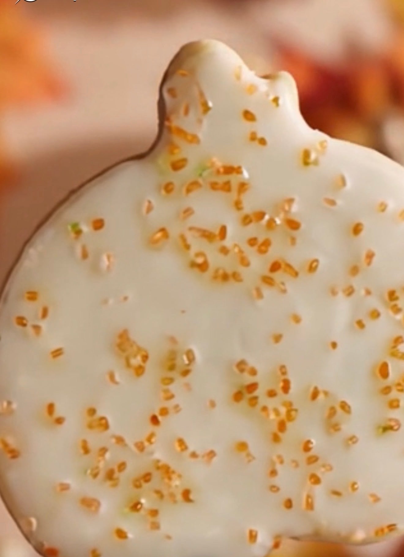 A close-up of a pumpkin-shaped cookie or candy coated in glossy white icing and sprinkled with gold sugar crystals, set against a warm, blurred background.