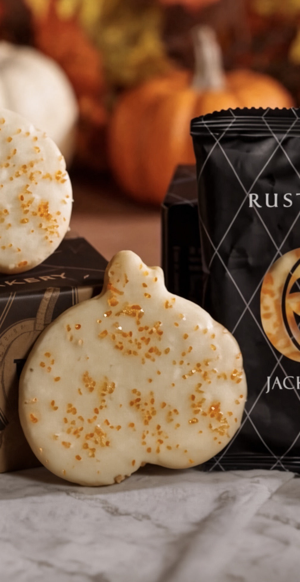A pumpkin-shaped white cookie with orange sprinkles in the foreground, with a black bag of Rustic Bakery  Cookies and more cookies blurred in the background among autumn pumpkins.