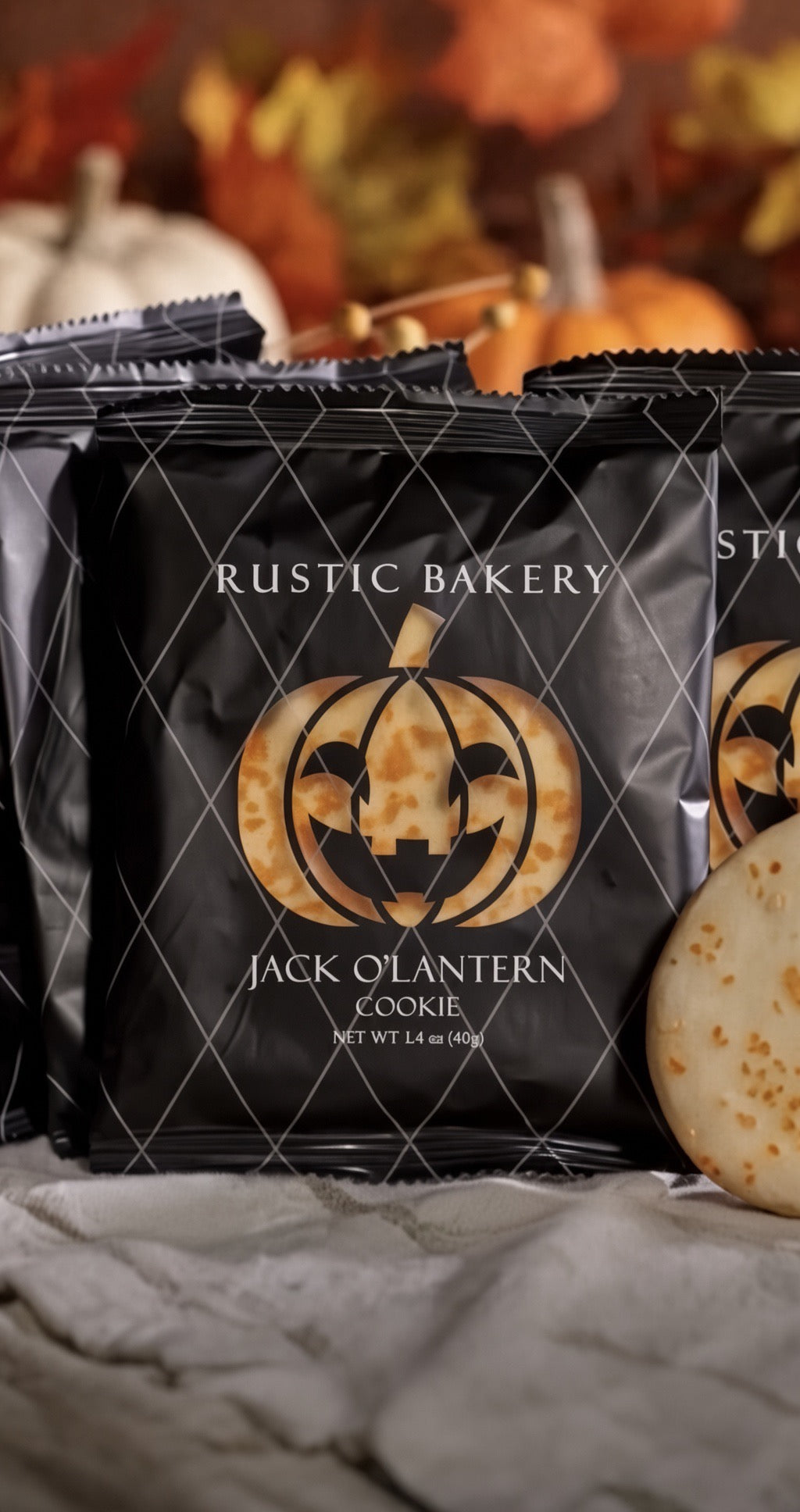 Black Rustic Bakery Jack O'Lantern Cookie bags arranged on a gray cloth with autumn pumpkins blurred in the background. A cookie is partially visible on the right side.