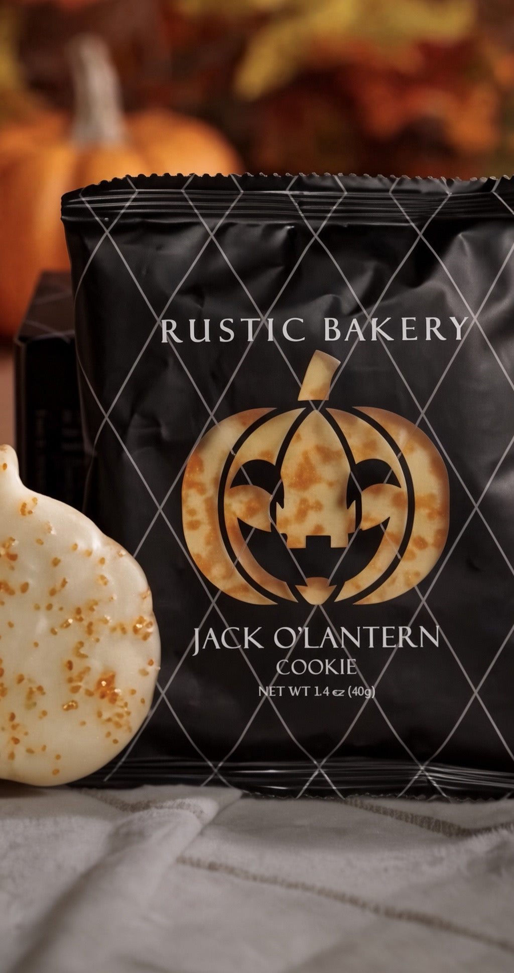 A black bag of Rustic Bakery Jack O'Lantern Cookie with a pumpkin graphic, shown next to a white cookie with gold sprinkles on a neutral fabric surface with a blurred autumn background.