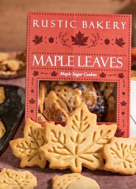 Rustic Bakery Maple Leaves maple sugar cookies in a red box, with several leaf-shaped golden-brown cookies displayed in front on a tabletop.