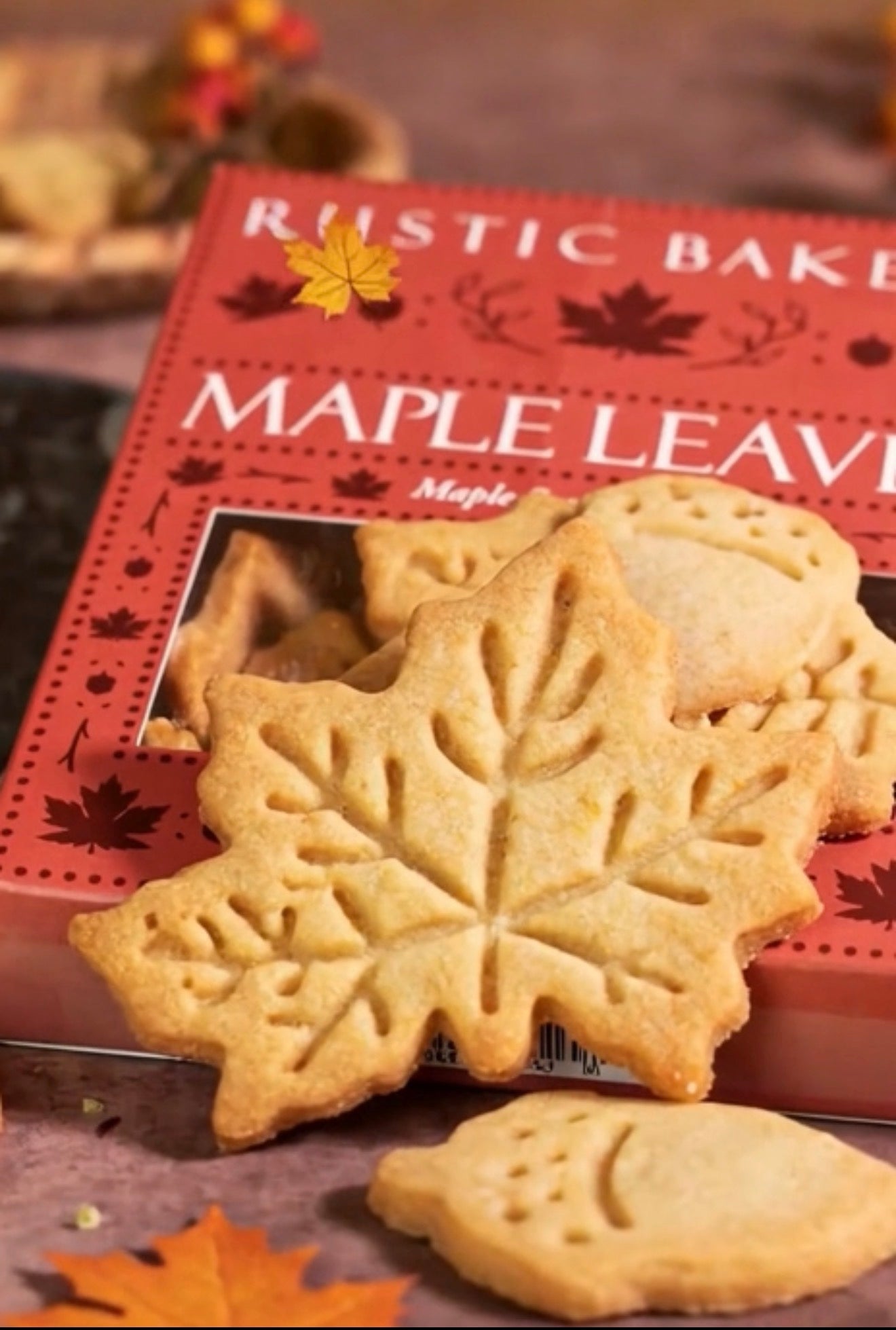 Rustic Bakery Maple Leaf cookies in front of their red packaging box, styled with autumn leaves on a rustic wooden surface.