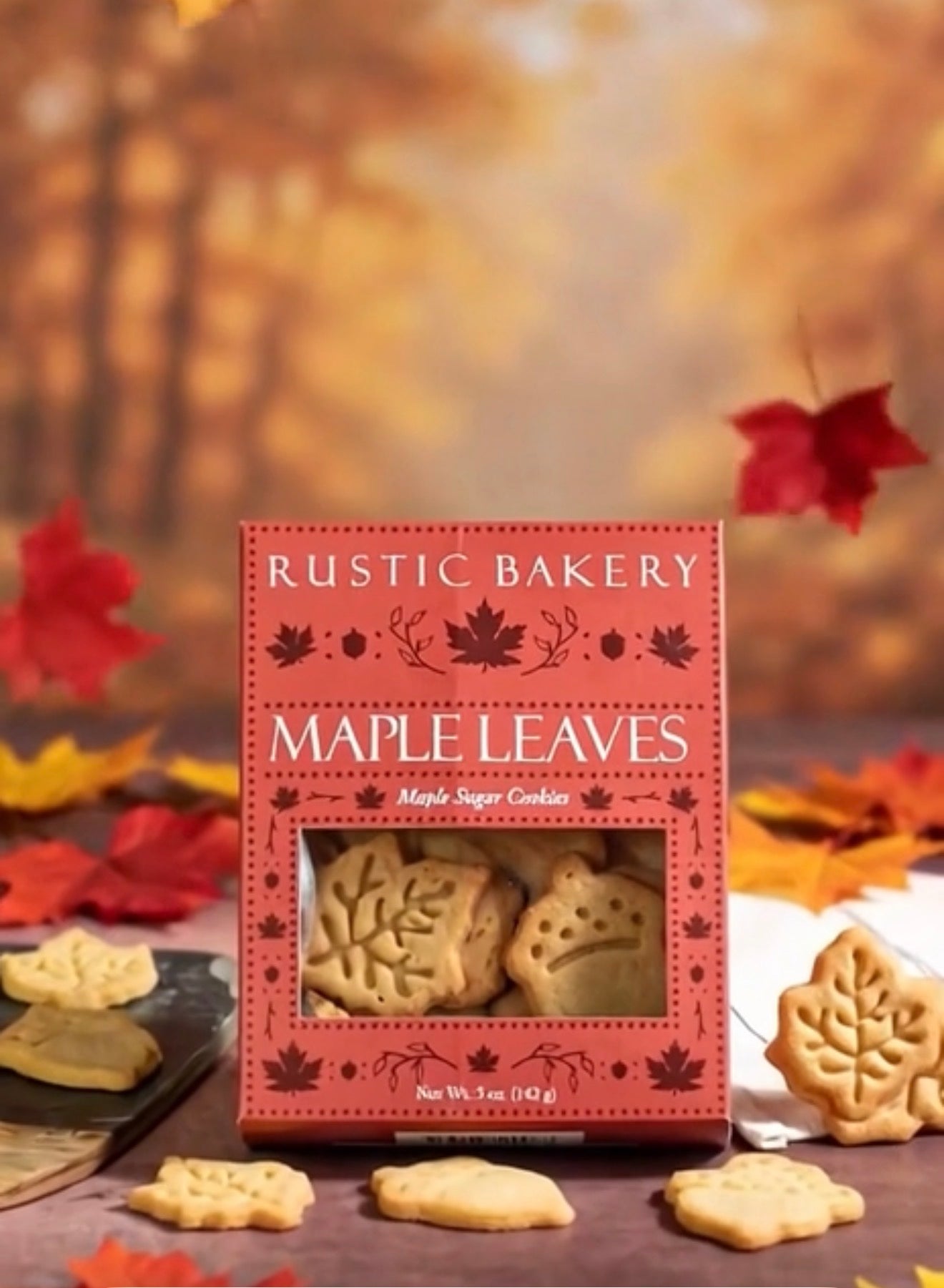 Rustic Bakery Maple Leaves maple sugar cookies in a red box, surrounded by scattered maple leaves and several leaf-shaped cookies on a rustic wooden surface with a warm autumn background.
