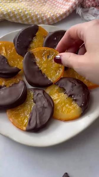 A hand with dark red nail polish reaches in to pick up a slice of orange half-dipped in chocolate from a white plate. More orange slices and chocolate are arranged on the plate, with a soft-focus kitchen background showing a checkered cloth.