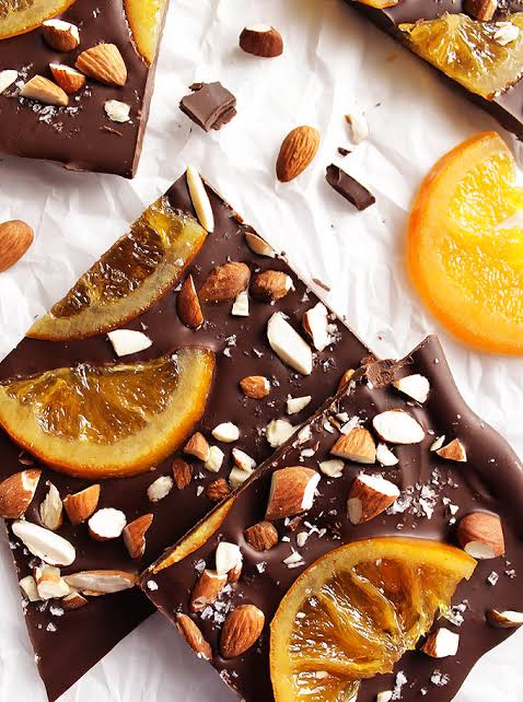 Chocolate bark pieces topped with dried orange slices, chopped almonds, and coarse sea salt, arranged on a white surface with scattered almonds and orange slices.