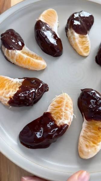 Several peeled orange segments dipped in glossy dark chocolate, arranged on a light gray plate.