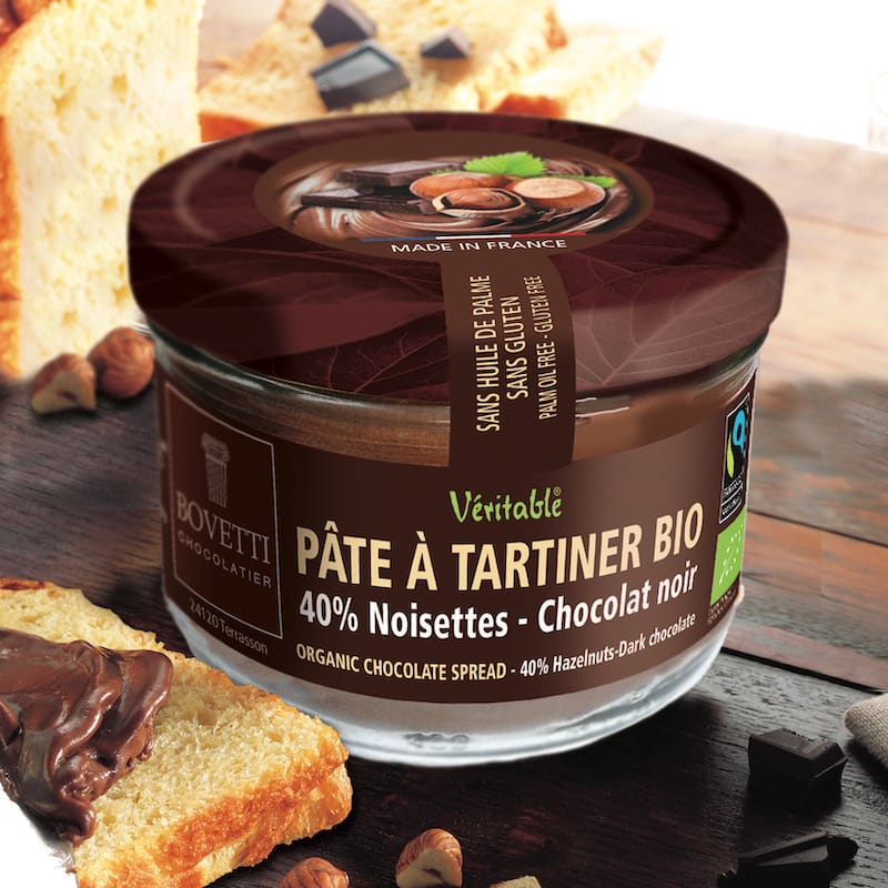 A jar of Bovetti Chocolater Bio organic chocolate spread (pâte à tartiner) with hazelnuts and dark chocolate pieces, styled on a rustic wooden table with bread slices.