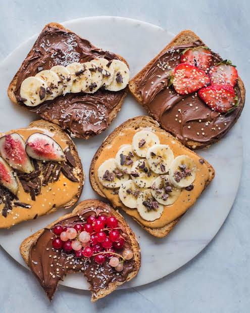 Five slices of toasted bread spread with various nut butters and fruit toppings, arranged on a round white plate against a light gray background.