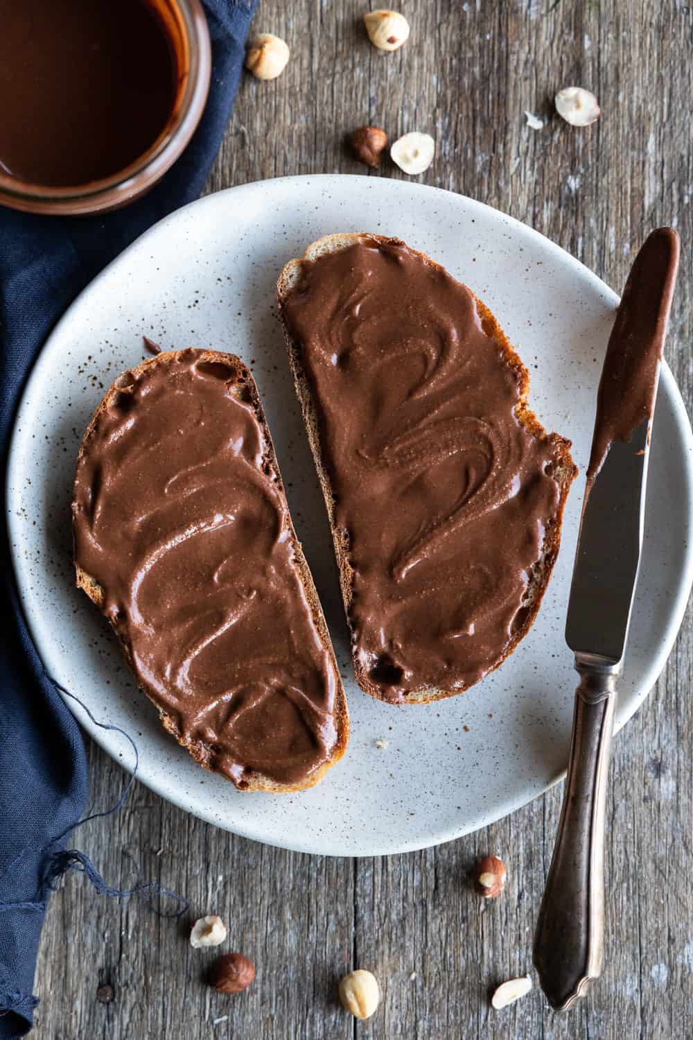 Two slices of bread spread thickly with chocolate hazelnut butter on a speckled white plate, set on a rustic wooden table with scattered hazelnuts, a knife, and a dark cloth.