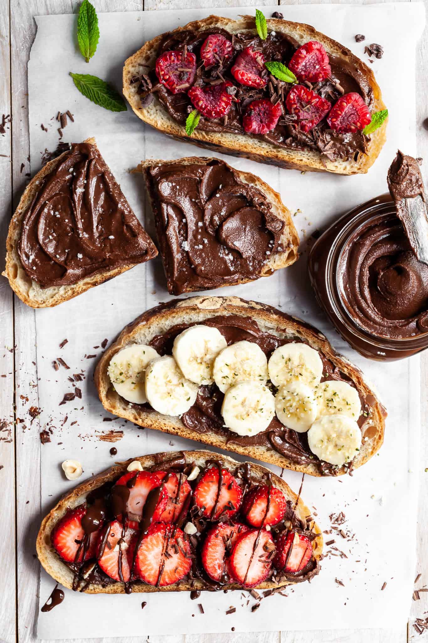 Four slices of toasted bread spread with chocolate hazelnut paste, topped with strawberries, bananas, or mint, arranged on a white wooden surface with a jar of chocolate spread nearby.