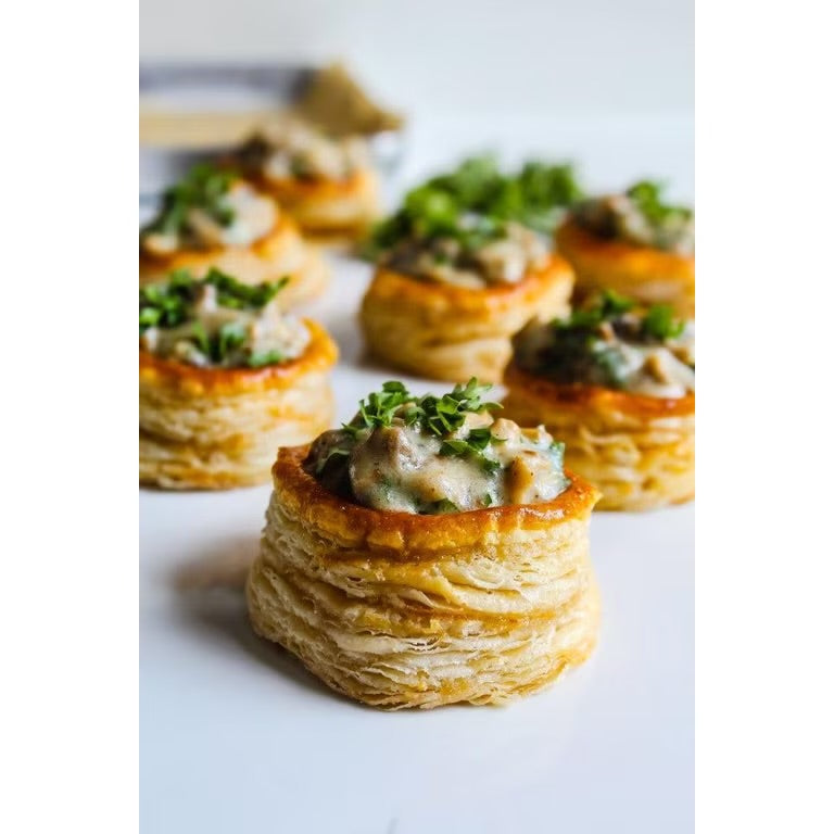 A close-up shot of several small flaky puff pastry cups filled with a mushroom and herb spread, garnished with chopped green herbs, arranged on a white surface.