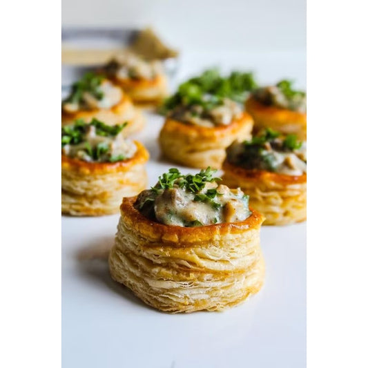 A close-up shot of several small flaky puff pastry cups filled with a mushroom and herb spread, garnished with chopped green herbs, arranged on a white surface.