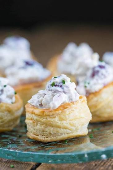 Close-up of several small puff pastry cups filled with creamy white appetizer mixture topped with purple bits and chopped green herbs, arranged on a glass plate.