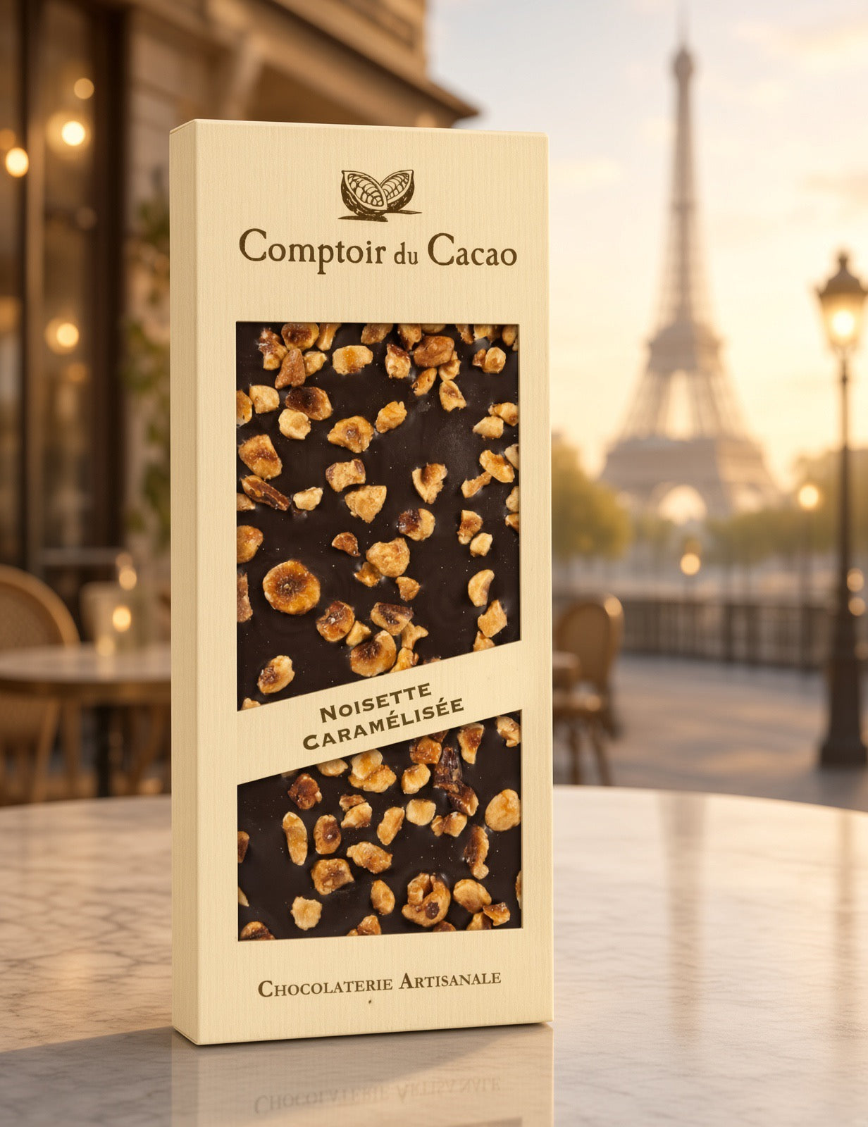 A tall cream-colored chocolate box labeled 'Comptoir du Cacao' and 'Noisette Caramélisée' stands on a marble table with a blurred Parisian street scene and Eiffel Tower in the background.
