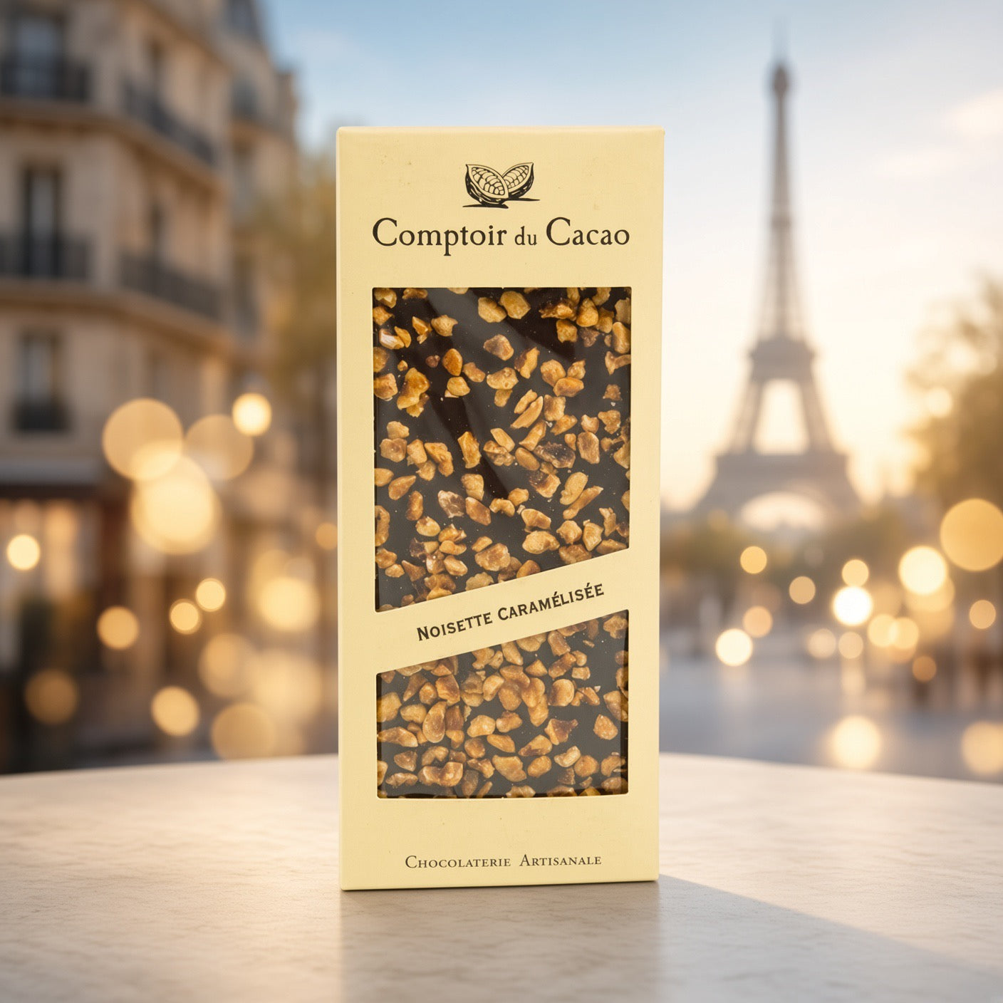 A vertical cream-colored box of Comptoir du Cacao Noisette Caramélisée chocolate, featuring two clear windows showing the dark chocolate with chopped nuts. The box is set on a light stone surface with a beautifully blurred Parisian street scene in the background, featuring the Eiffel Tower and warm bokeh lights.
