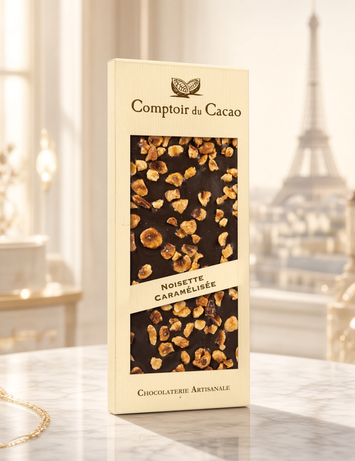 A package of Comptoir du Cacao Noisette Caramélisée chocolate stands on a marble surface with a softly blurred background showing a bright interior and the Eiffel Tower.