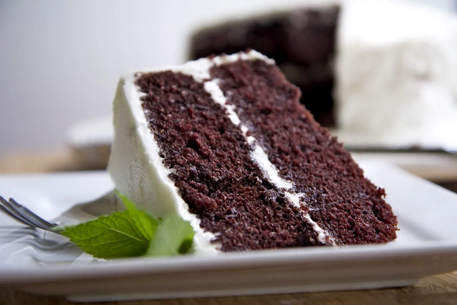 A slice of two-layer chocolate cake with white frosting on a white plate, garnished with a green leaf, with a blurred whole cake in the background.