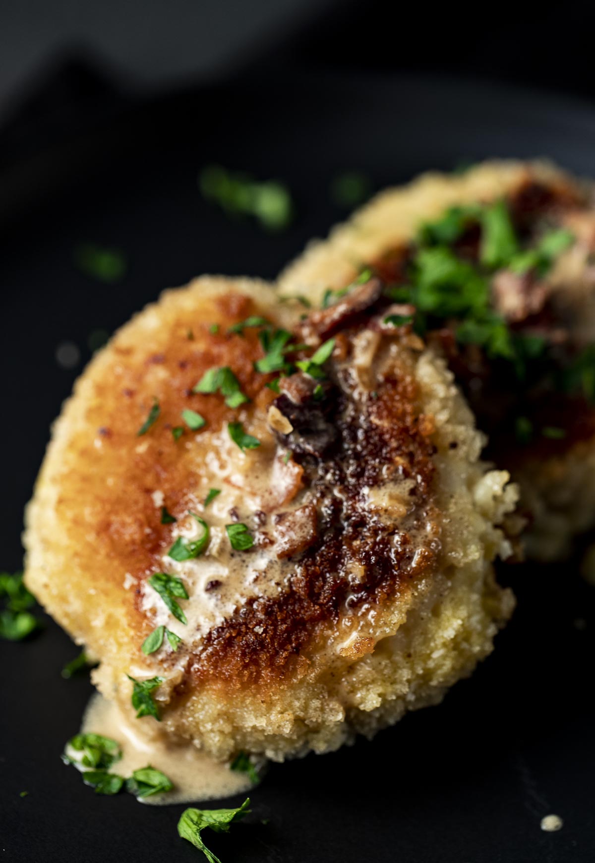 Two golden-brown fried breaded cutlets topped with a creamy sauce and chopped green herbs, served on a dark plate.