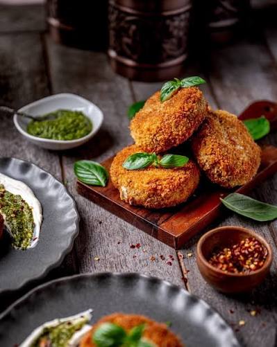 Three golden-brown breaded cutlets garnished with fresh basil leaves, served on a wooden board with small bowls of green sauce and red pepper flakes on a rustic wooden table.