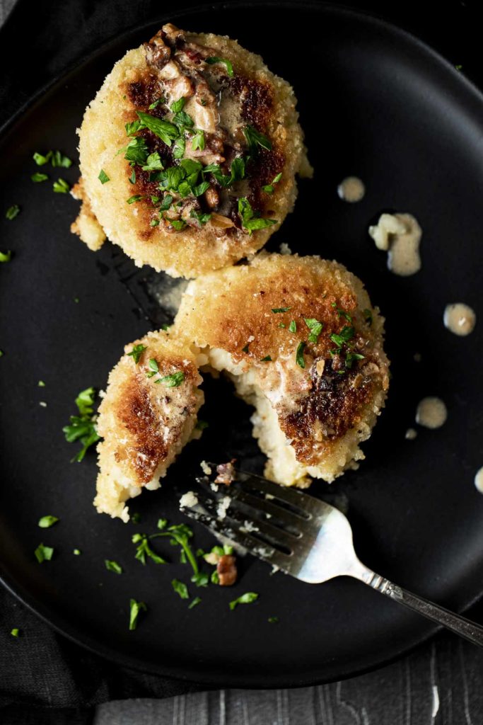 Two golden-brown breaded patties topped with chopped green herbs and mushrooms, served on a black plate with a fork and a drizzle of sauce.
