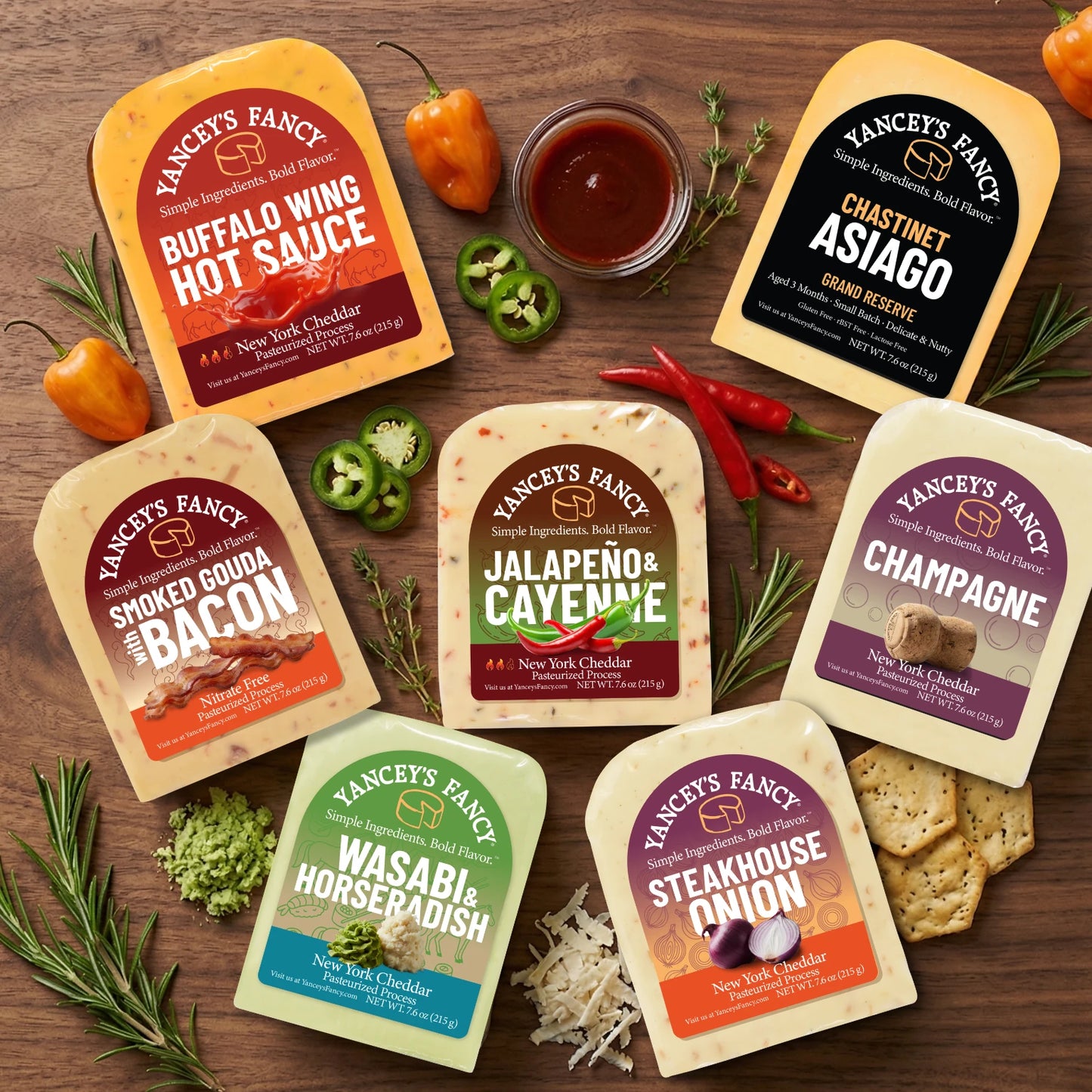Seven packaged snack and sauce products from Yancey's Fancy are arranged on a wooden surface with food props including sliced jalapeño, chilis, herbs, crackers, and a small bowl of sauce.