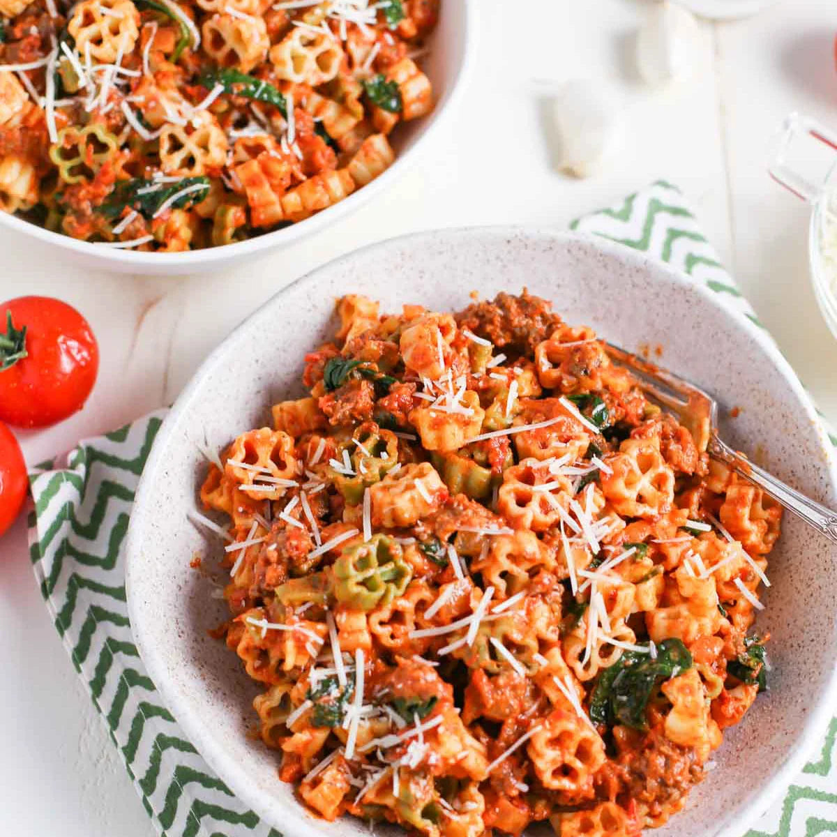A bowl of pasta meat and sausage in a rich tomato sauce, topped with shredded cheese and green vegetables, served on a white table with a green patterned napkin and a second bowl in the background.