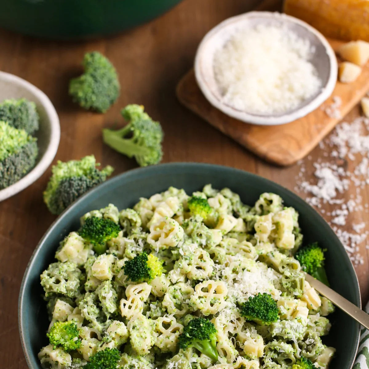 A bowl of broccoli pasta with a creamy green sauce, garnished with grated cheese, on a wooden table with broccoli florets and a small bowl of cheese nearby.